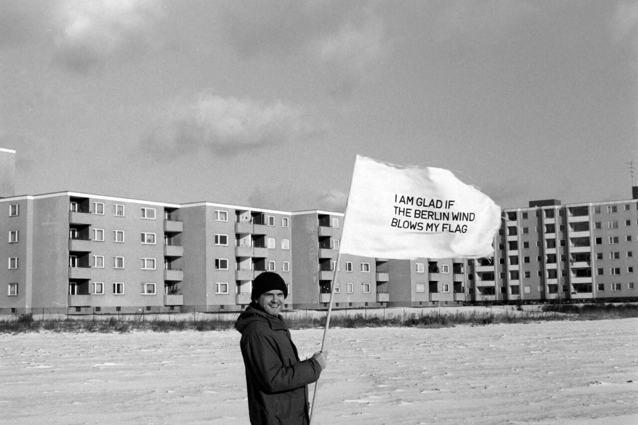 Eine Person hält eine Flagge mit der Aufschrift "ICH FREUE MICH, WENN DER WIND MEINE FLAGGE WEHT" vor einem Wohngebäude.