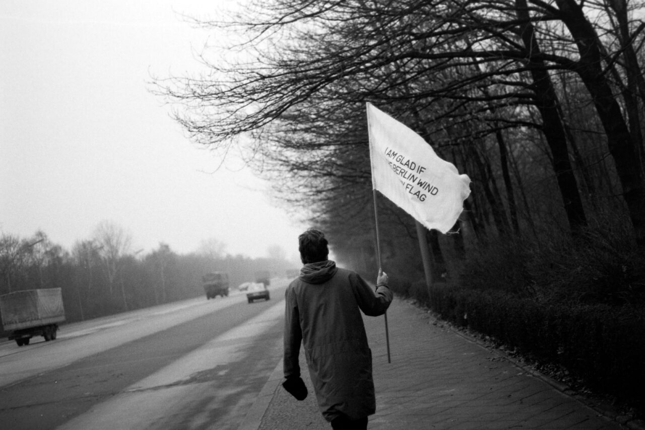 Eine Person hält eine Flagge und läuft an einer Straße entlang, während im Hintergrund Bäume zu sehen sind. Schwarz-weißes Bild.
