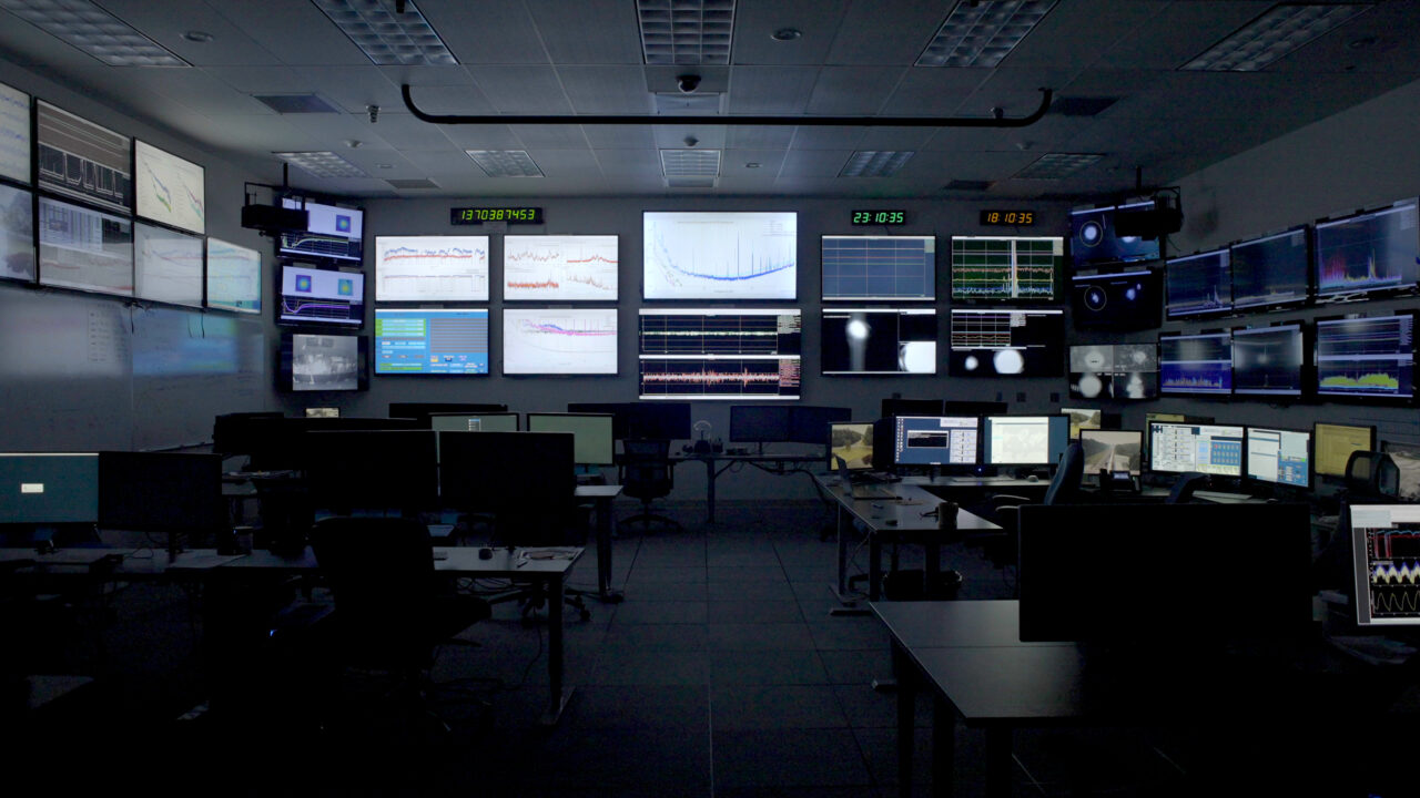 Moderner Kontrollraum mit mehreren Bildschirmen, die Daten und Grafiken in einer dunklen Umgebung anzeigen.