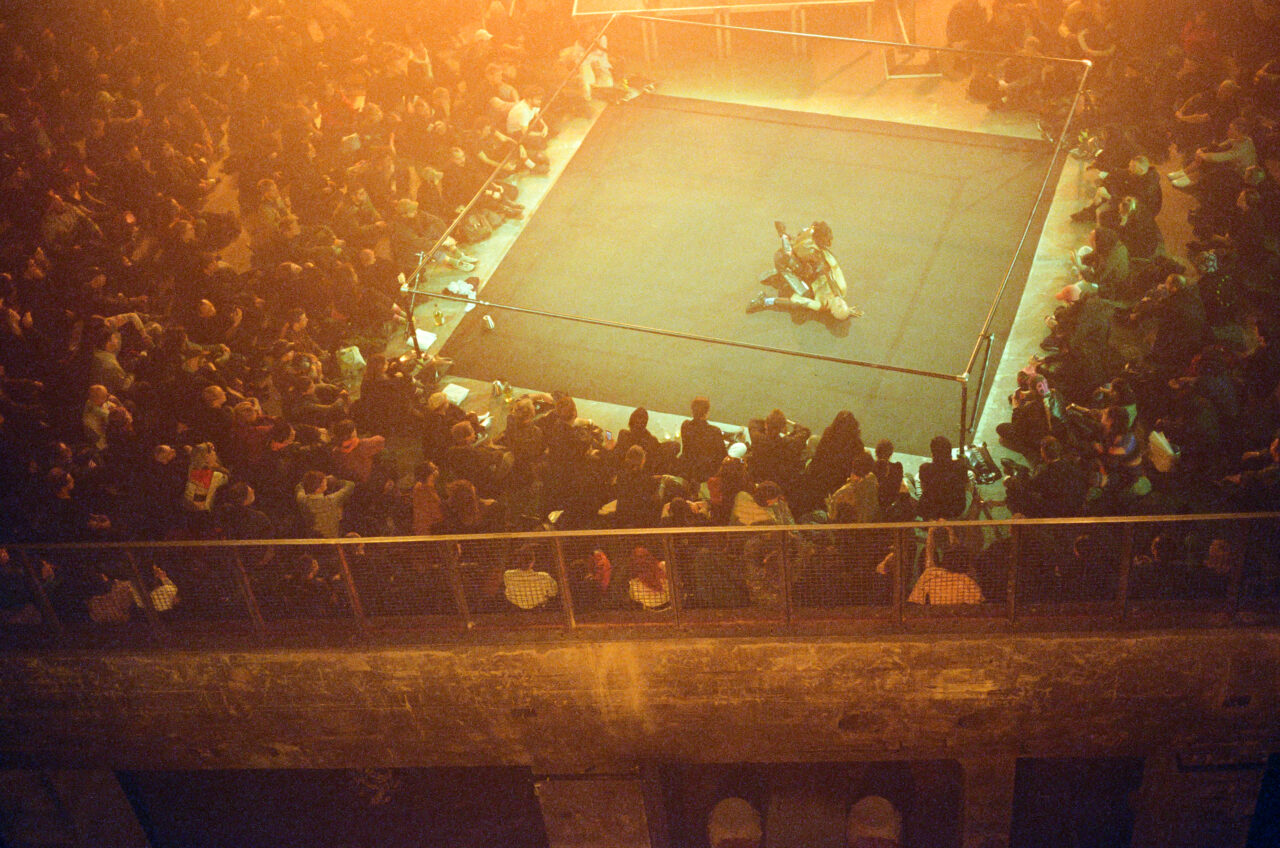 Overview of a packed room with a ring in the center where two fighters face off.