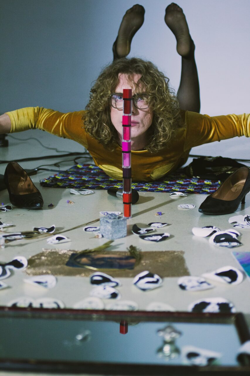 A woman with curly hair lies on the floor, surrounded by mirrors and colorful objects, wearing a yellow top.
