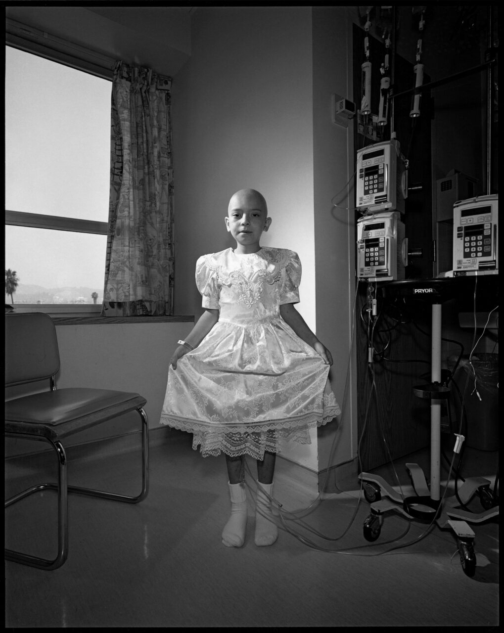 Ein Mädchen in einem weißen Kleid steht neben medizinischen Geräten in einem Krankenhauszimmer.