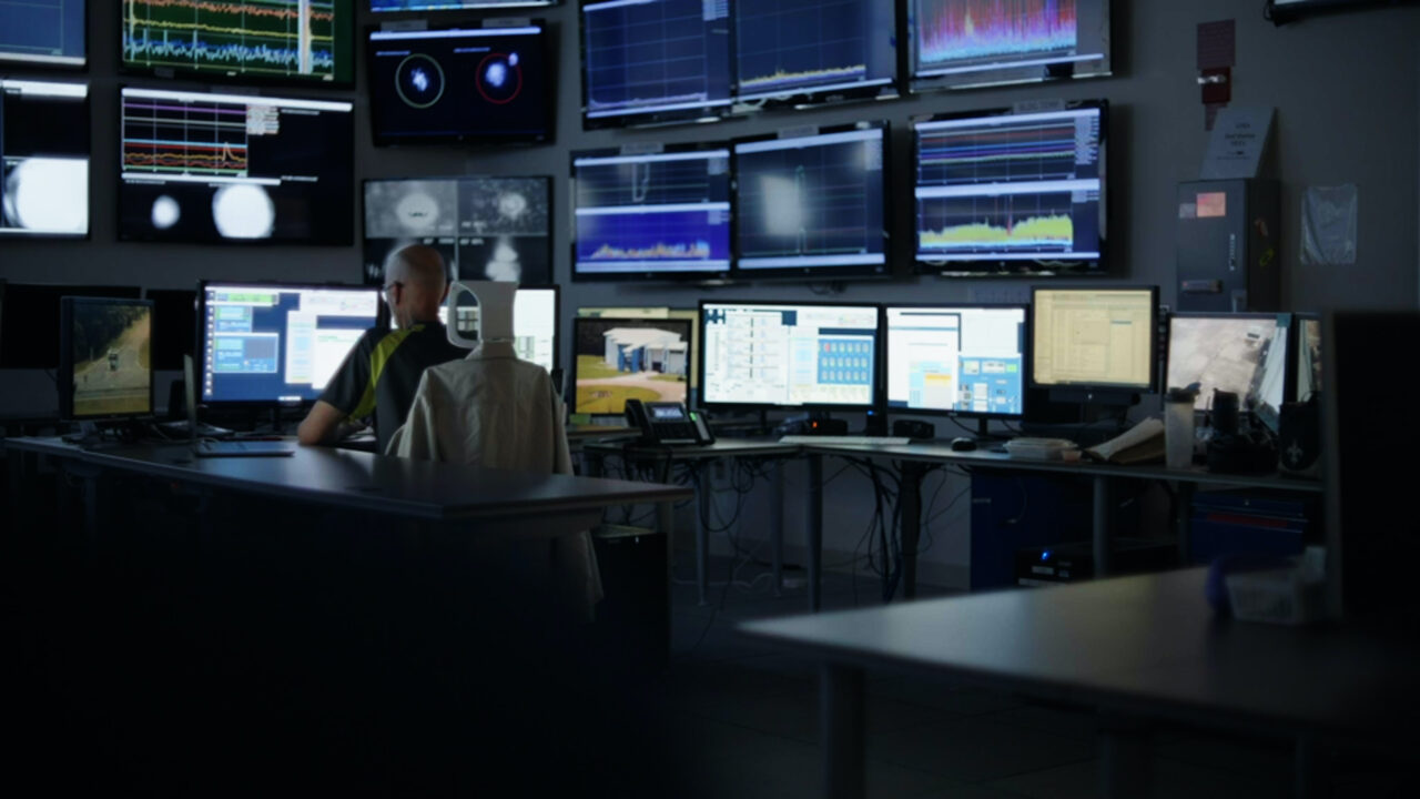 Person in einem Kontrollraum mit mehreren Bildschirmen, die Daten und Analysen darstellen.