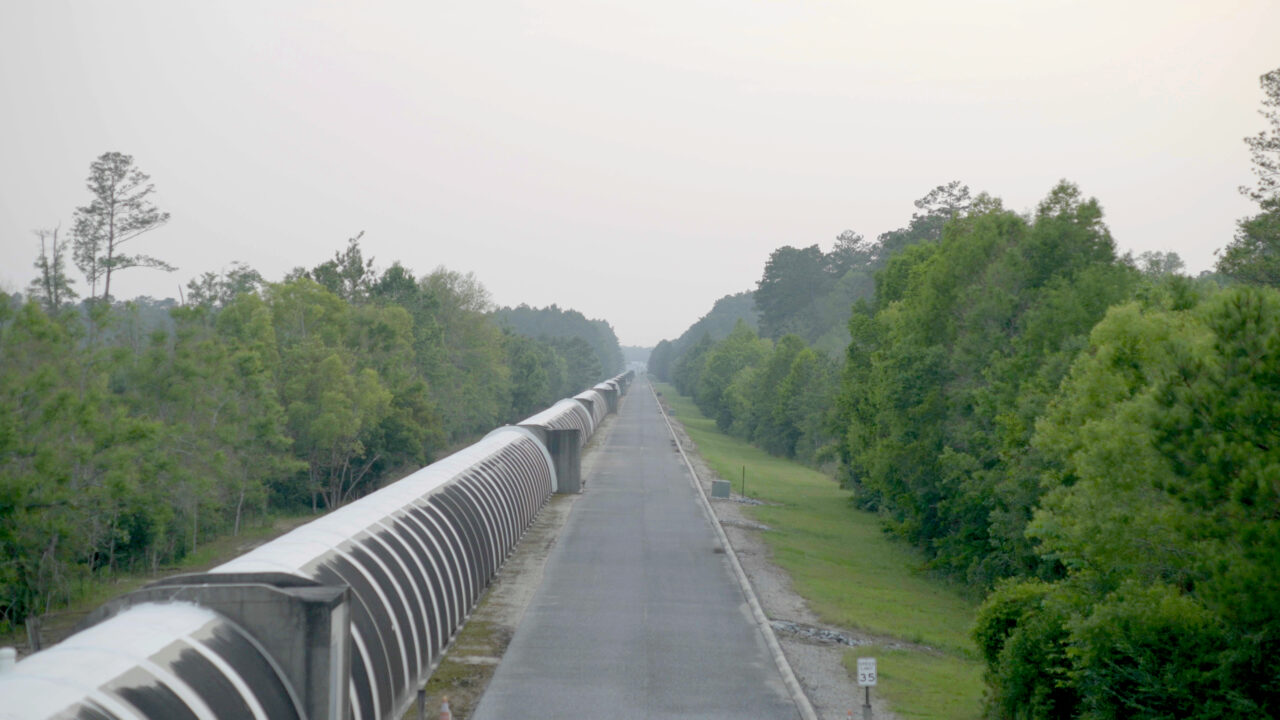 Gerader Weg mit Eisenbahnlinie, umgeben von Bäumen und sanften Hügeln unter einem nebligen Himmel.