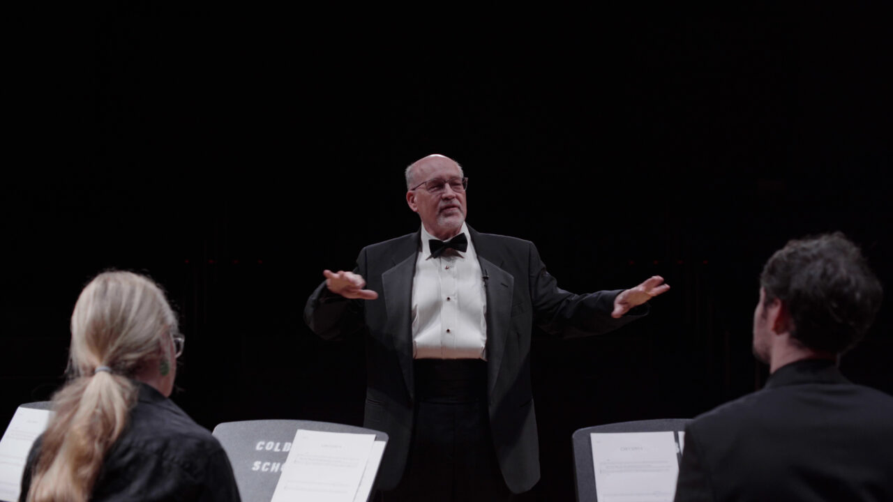 Der Dirigent leitet das Orchester, während die Musiker konzentriert auf ihre Notenständer blicken. Dunkler Hintergrund.