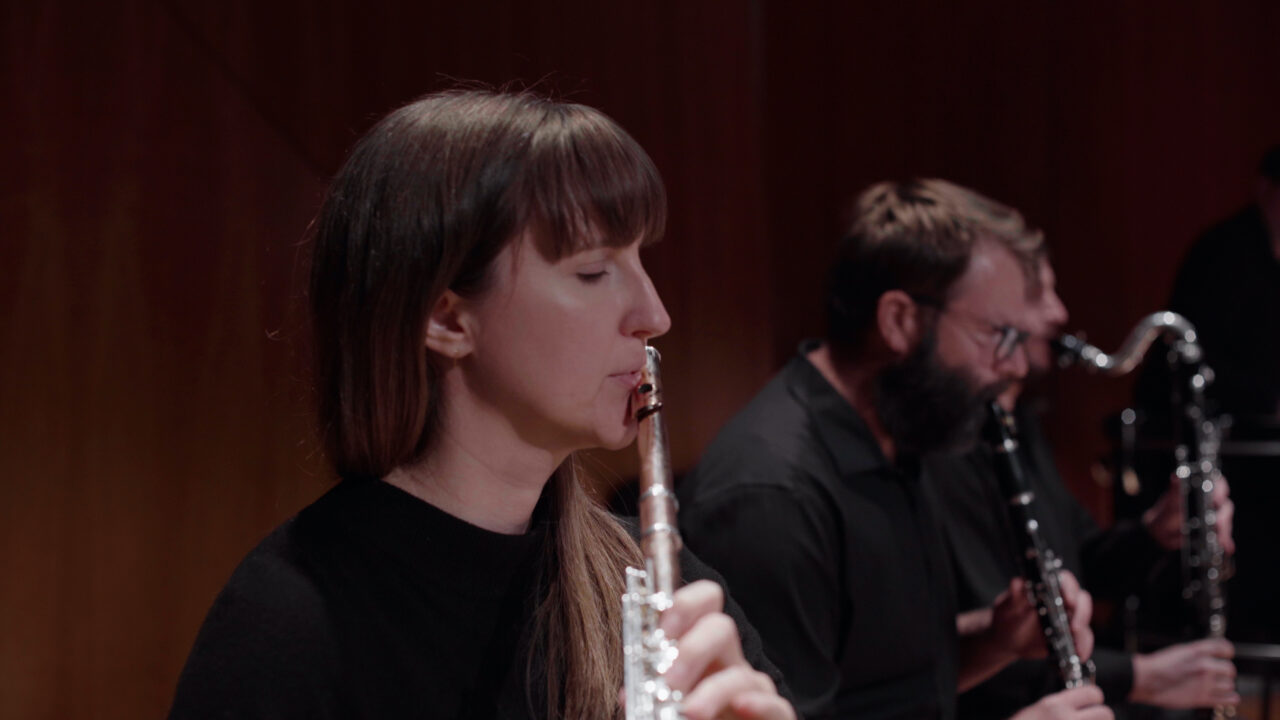 Musikerin spielt Flöte in einem Ensemble; im Hintergrund sind andere Musiker mit Klarinetten zu sehen.