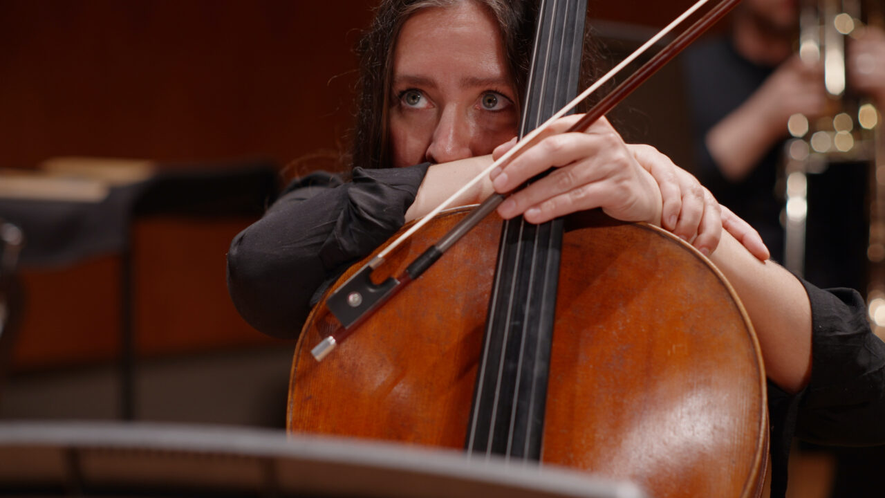 Frau mit Cello in einem Orchester, konzentriert beim Spielen, weitere Musiker im Hintergrund.