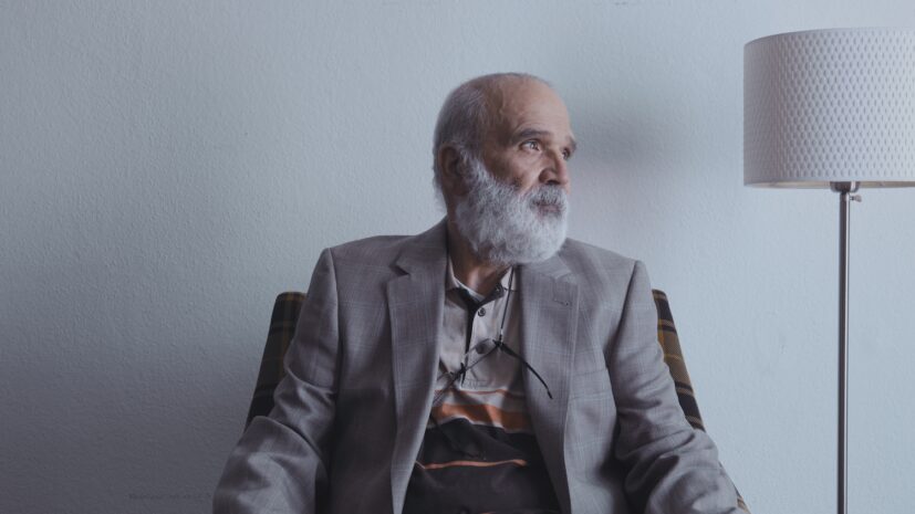 An older man with a gray beard sits in an armchair, thoughtfully looking to the side, with a table lamp beside him.