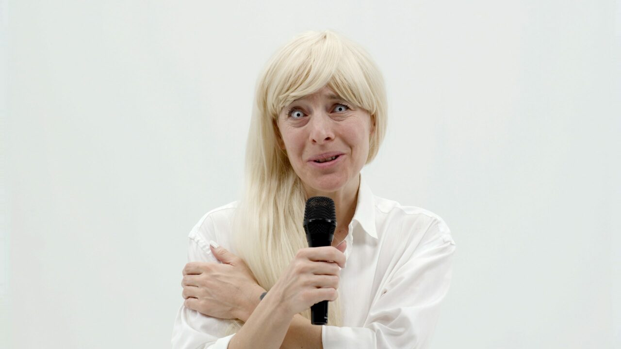 A woman with blonde hair holds a microphone, smiles, and speaks against a white background.