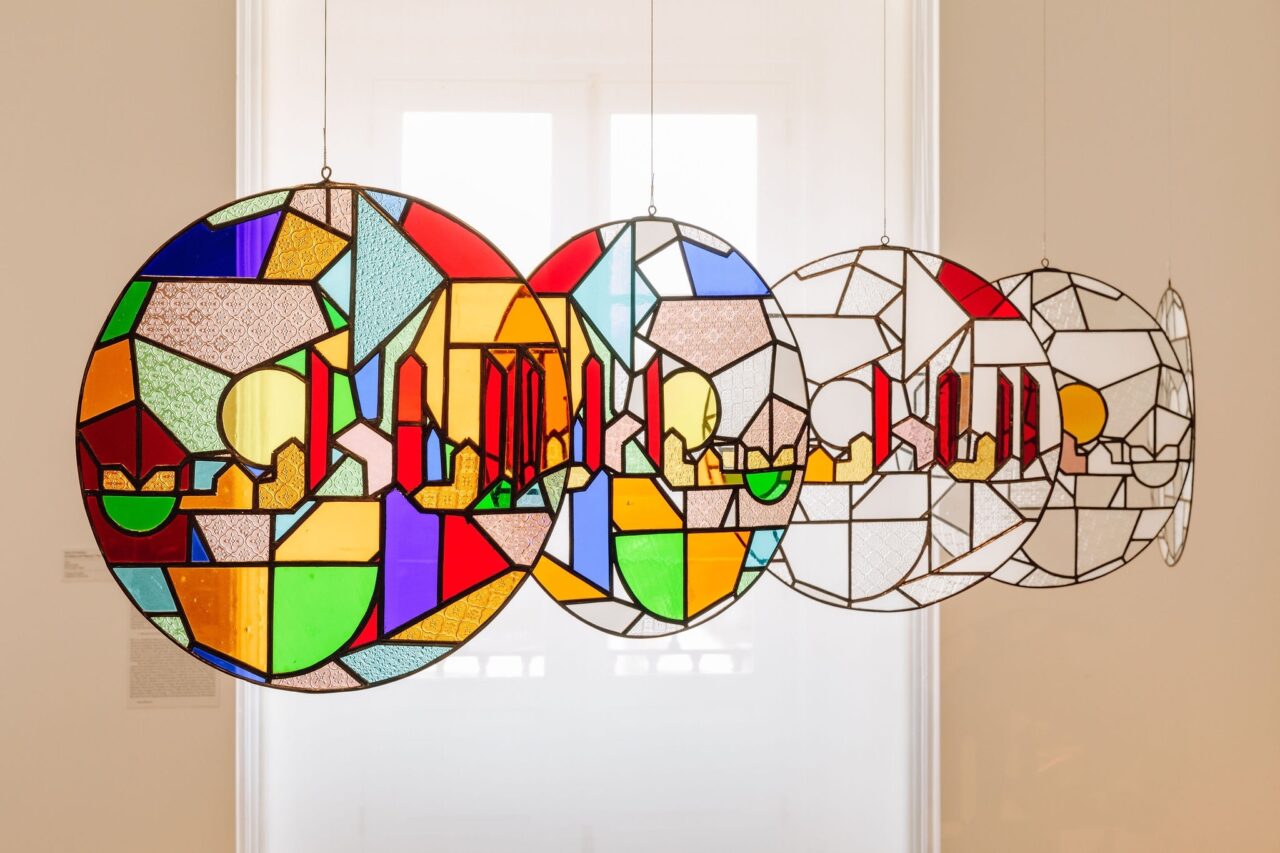 Buntes Glasfenster mit geometrischen Mustern in verschiedenen Farben, schwebend in einem hellen Raum.