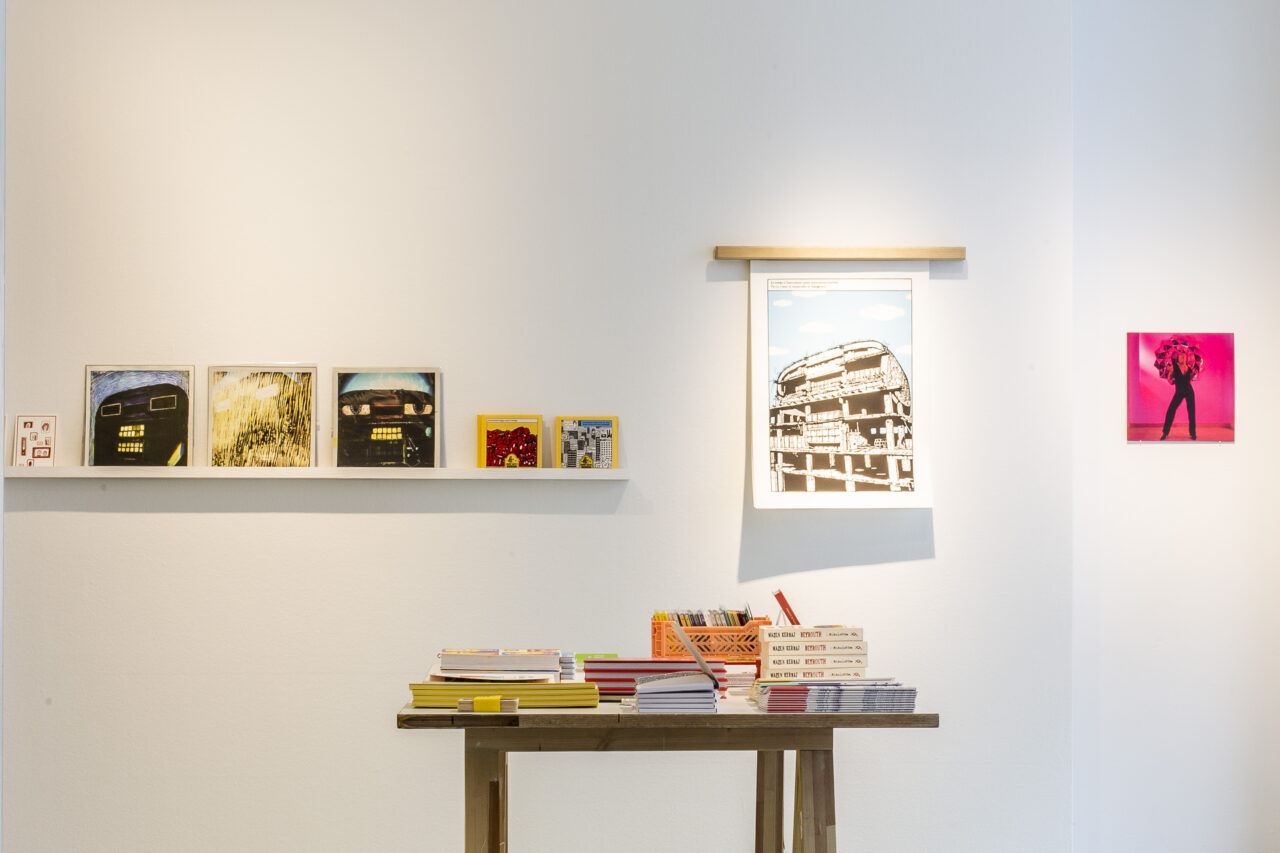 Ein moderner Ausstellungsraum mit Kunstwerken an der Wand und einem Tisch mit vielen Büchern und Zeitschriften.