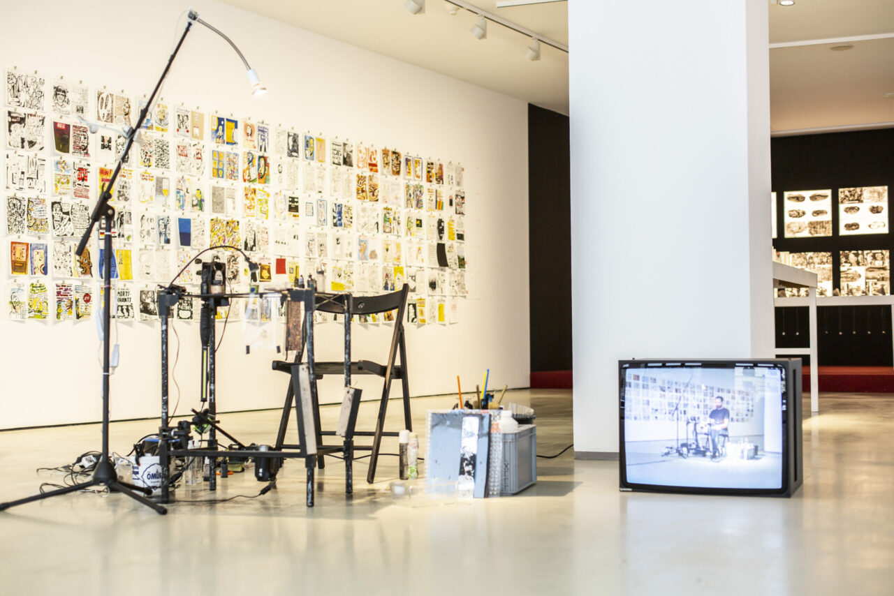 Artistic setup in a gallery with chairs, lighting, and an old television against a wall full of pictures.