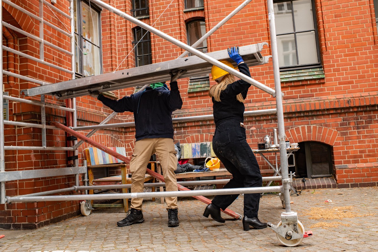 Two people are lifting a scaffold, with a red brick building in the background.