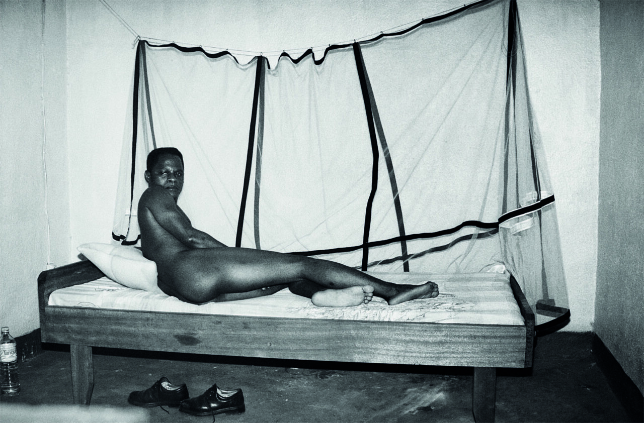 A person lies naked on a bed, surrounded by a mosquito net in a simple room.