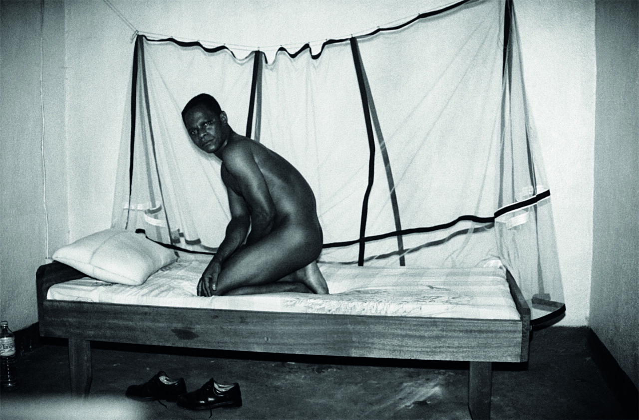 Naked person sits on a bed surrounded by a mosquito net in a simple room.