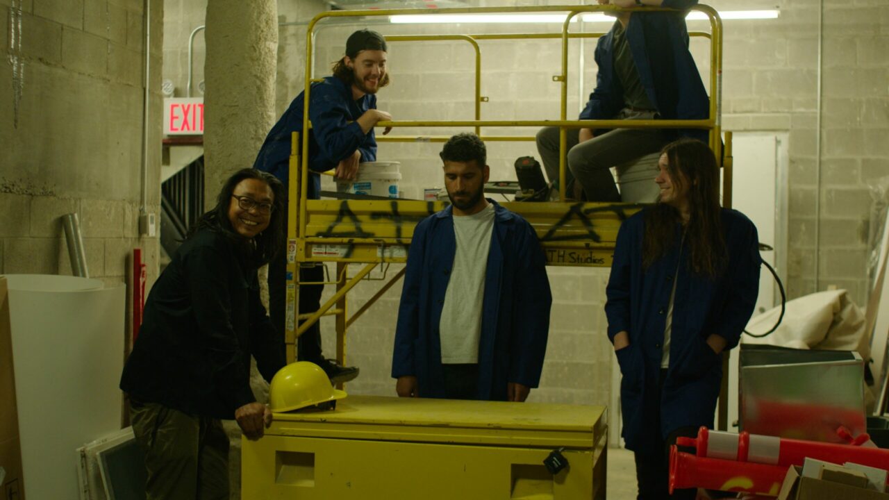 Four people in blue overalls stand in a storage room, two on a scaffold and two in front with a yellow container.