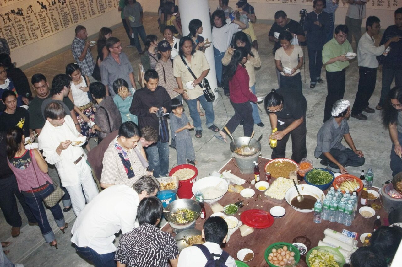 Eine Menschenmenge versammelt sich um einen Tisch mit Essen, während einige auf das Essen oder Fotos warten.