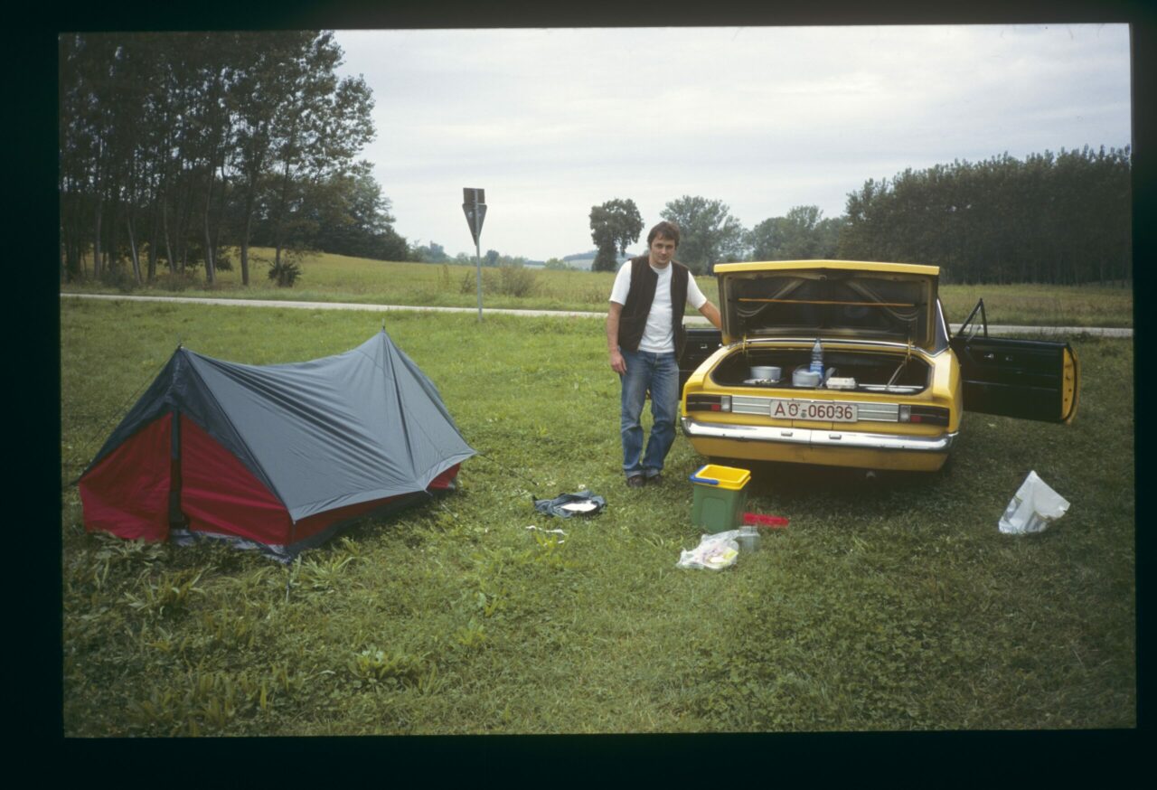 A man stands next to a yellow car, with a tent set up in the background on a meadow.