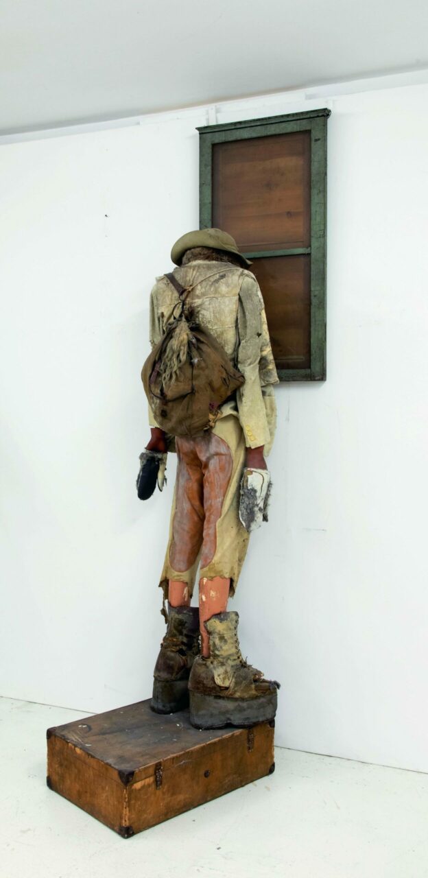 Sculpture of a standing man in worn clothing, with a backpack and hat, looking at a window.