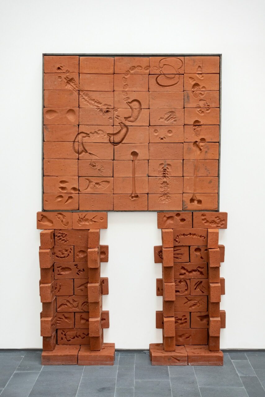 Red sculpture made of brick with relief patterns, consisting of a wall and two supporting columns.