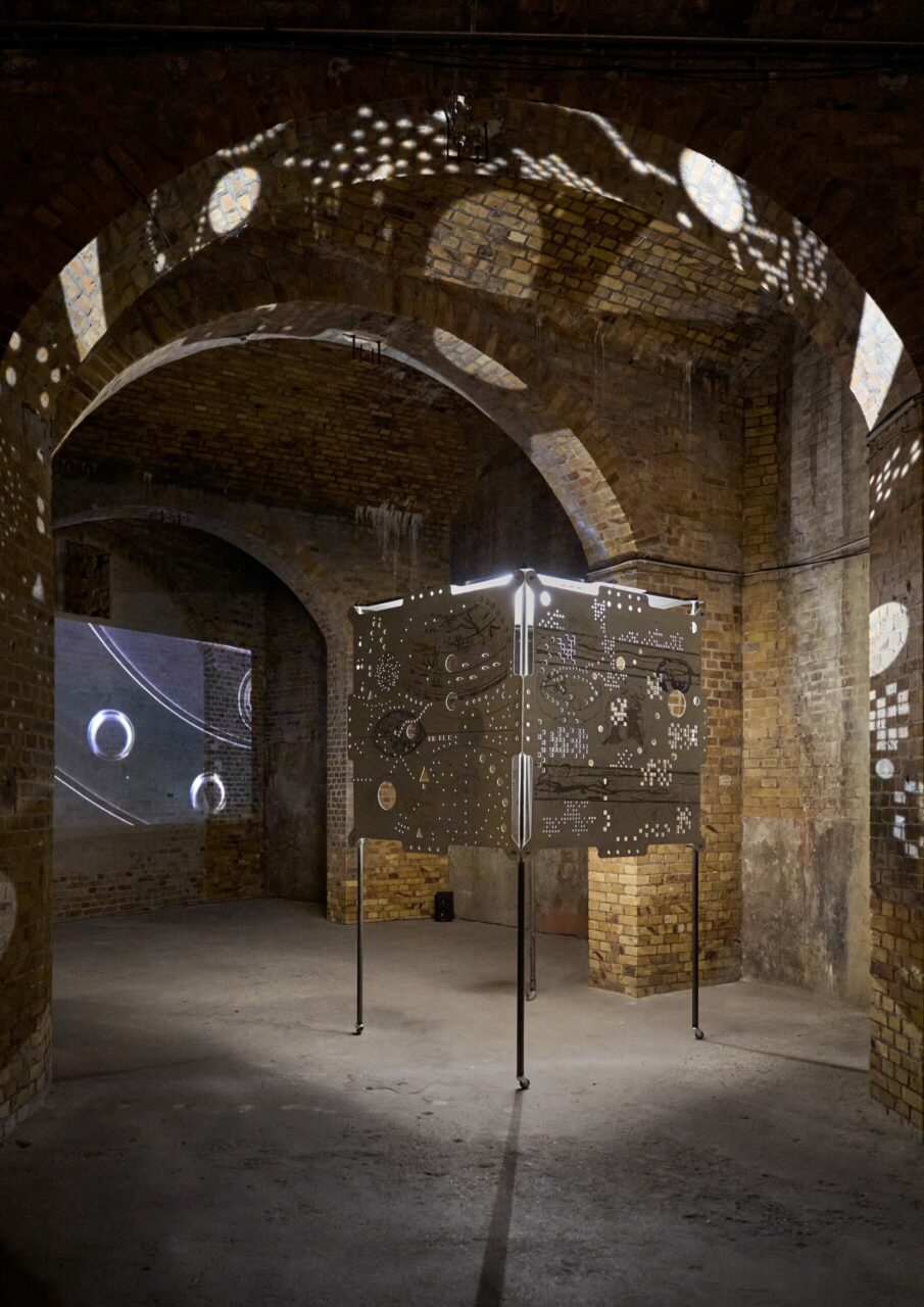 Art installation in a historic vault featuring a glowing, decorative cube and projected images on the walls.