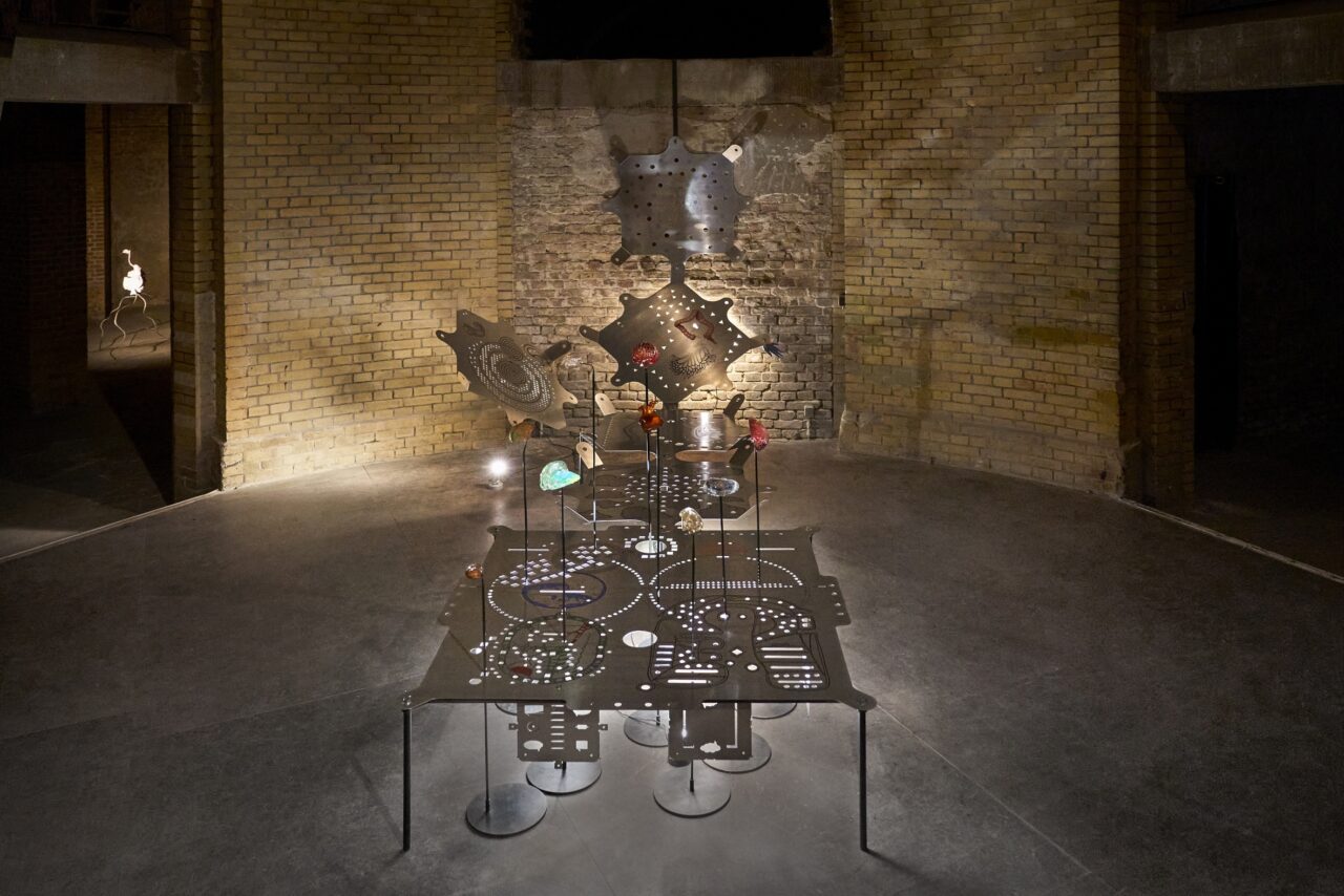Modern exhibition with a central table surrounded by artistic objects and a rough brick wall.