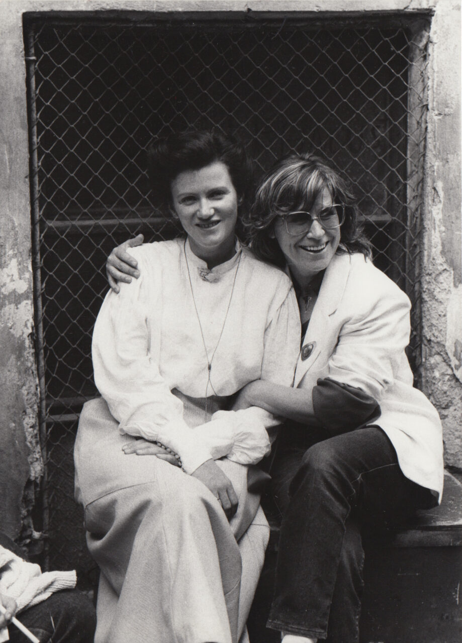 Zwei Frauen umarmen sich und lächeln in einer urbanen Umgebung, festgehalten in einer Schwarz-Weiß-Fotografie.