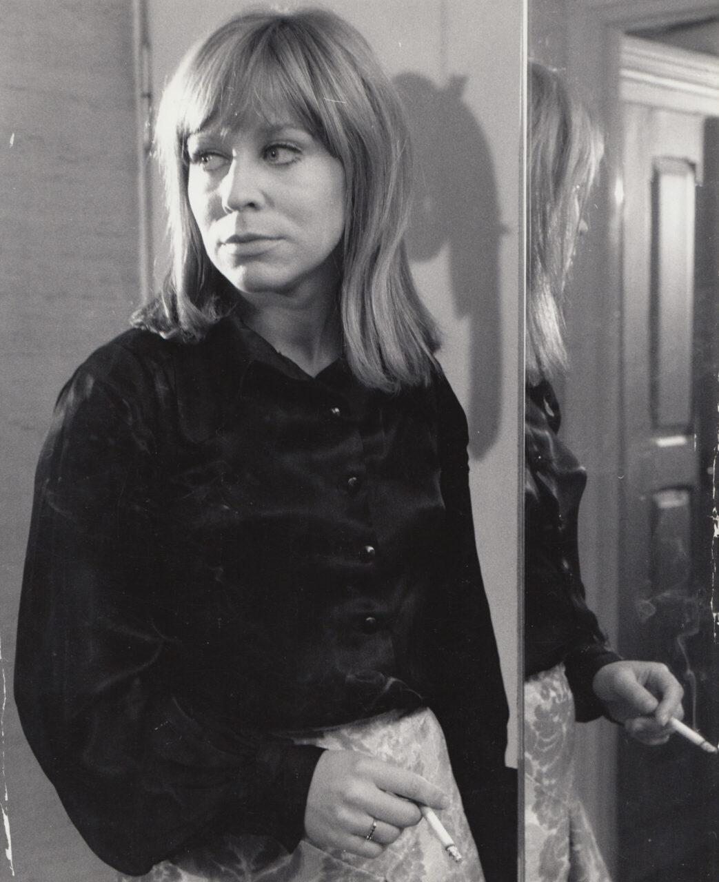 A woman with blonde hair in a black blouse gazes pensively into a mirror. Black and white photo.