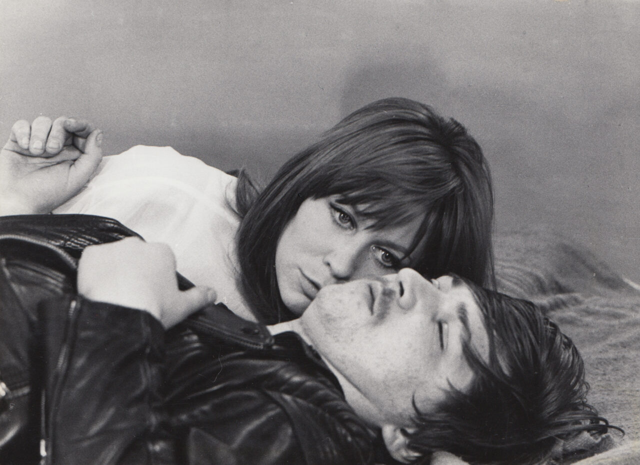 Ein Paar liegt auf einer Matratze, der Mann in Lederjacke, die Frau sieht ihn an und wirkt nachdenklich.