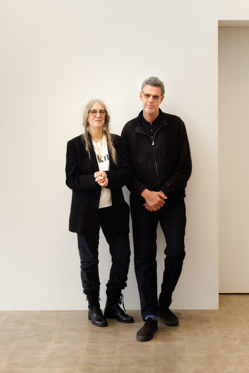 Zwei Personen in dunkler Kleidung stehen vor einer weißen Wand, beide mit neutralem Gesichtsausdruck.