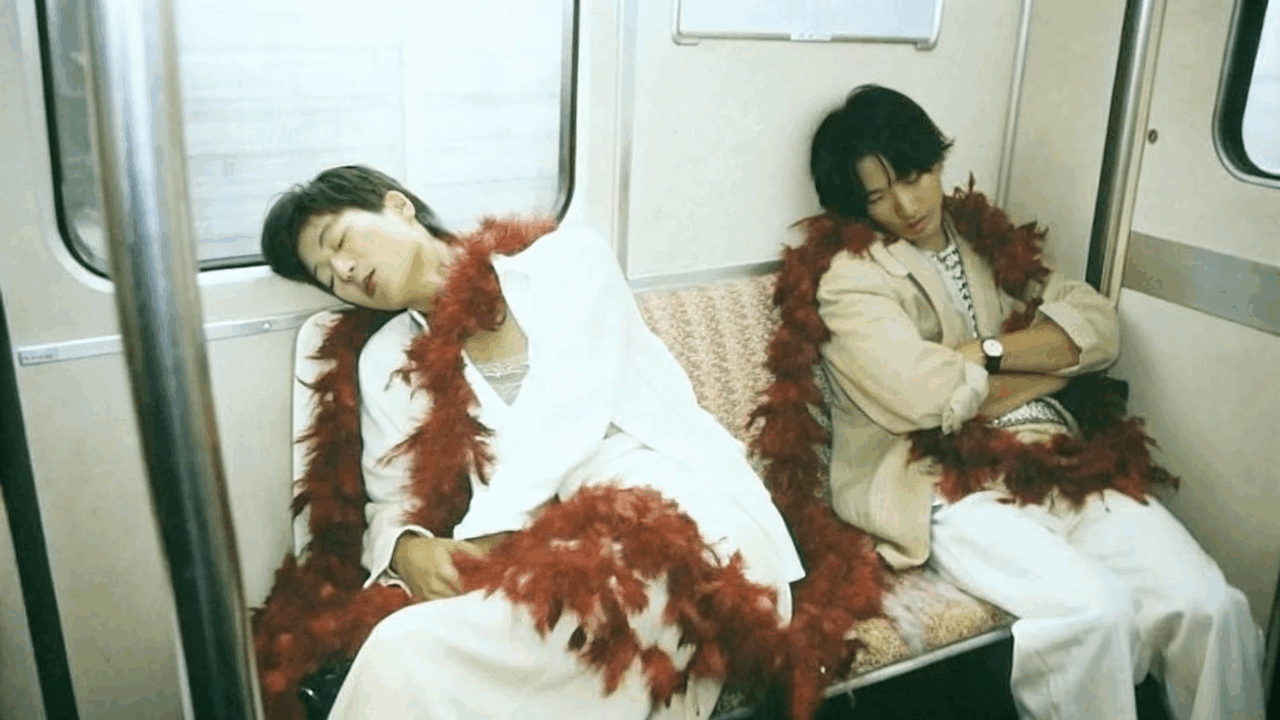 Zwei Personen schlafen in einer U-Bahn, beide mit roten Federboas, sitzen auf einer Bank.