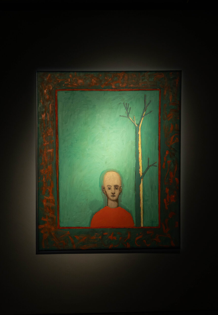 Portrait of a person with a shaved head, wearing a red top, with a tree on a green background, in a bright frame.