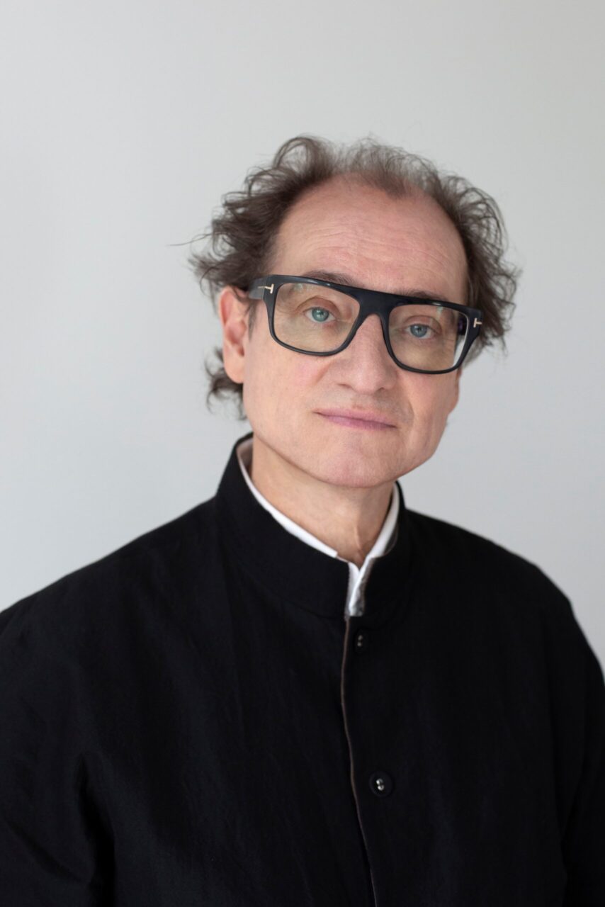 Portrait of a man with curly hair, glasses, and black clothing against a neutral background.