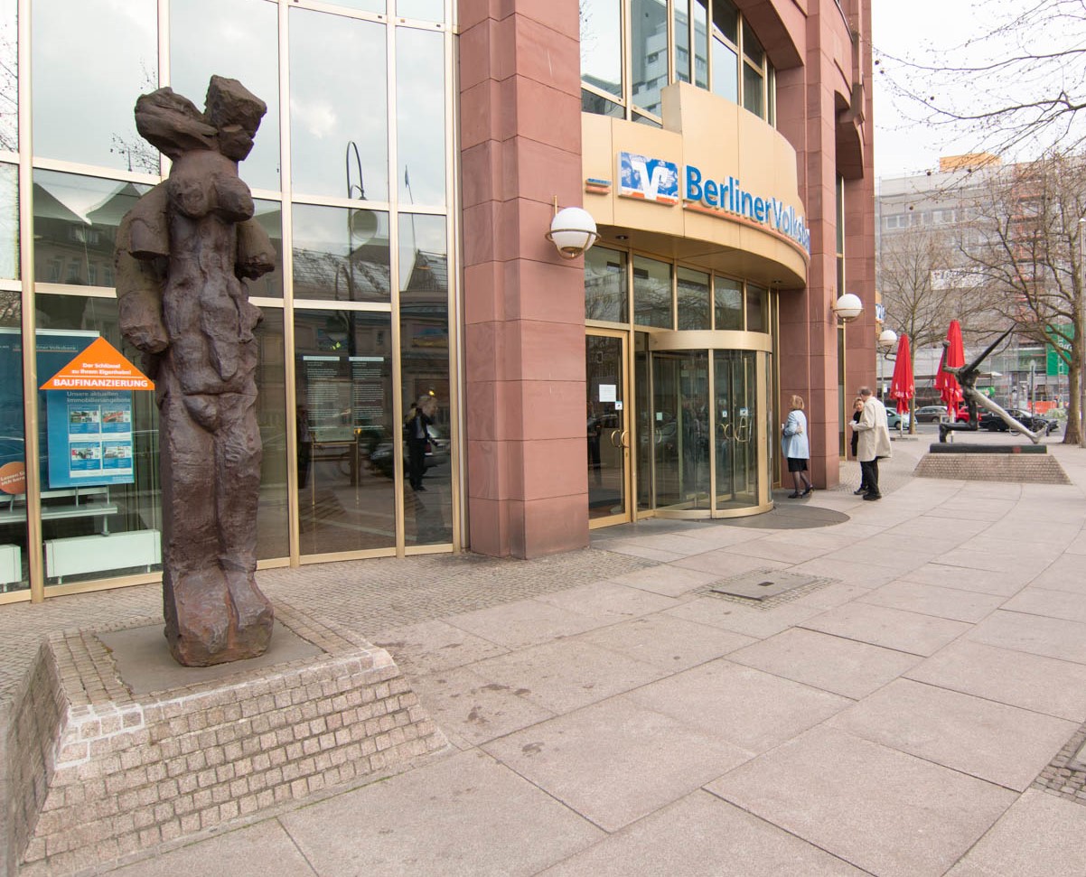 Eingang der Berliner Volksbank mit einer modernen Skulptur und Passanten, die draußen stehen.
