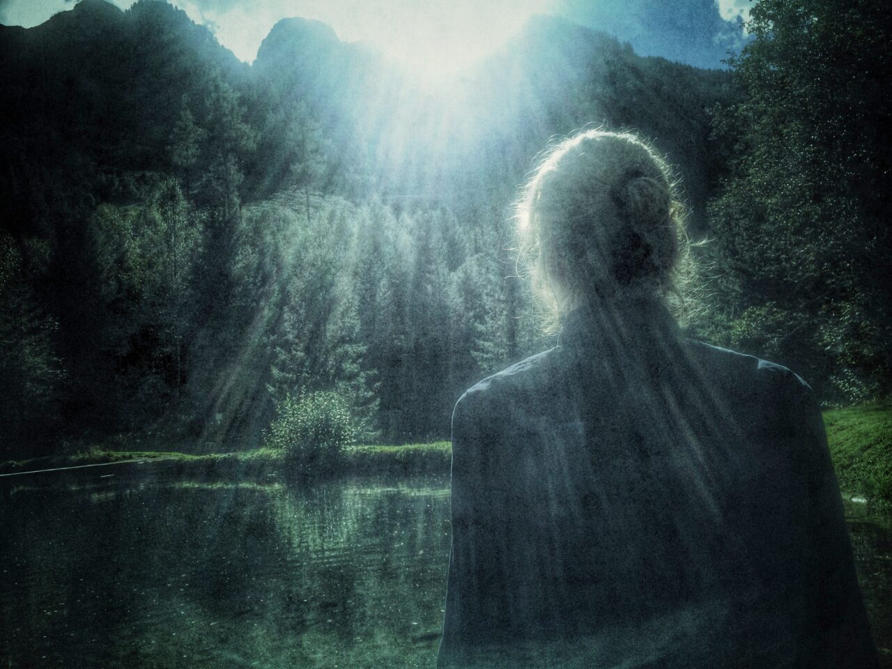 Person with their back to a lake, surrounded by mountains and trees, light rays breaking through the darkness.