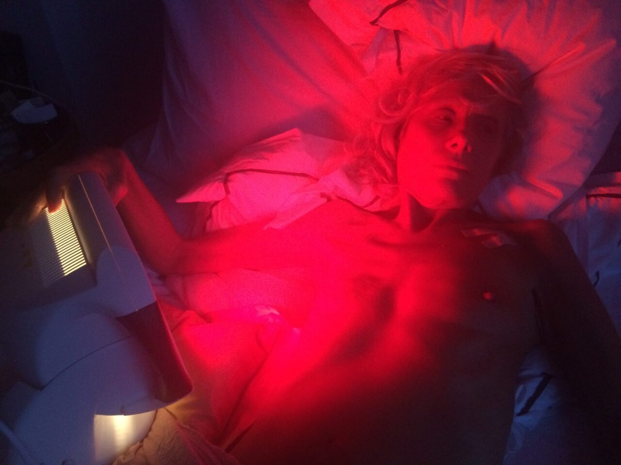 Person mit blonden Locken liegt im Bett und erhält eine rote Lichtbehandlung mit einem Gerät.