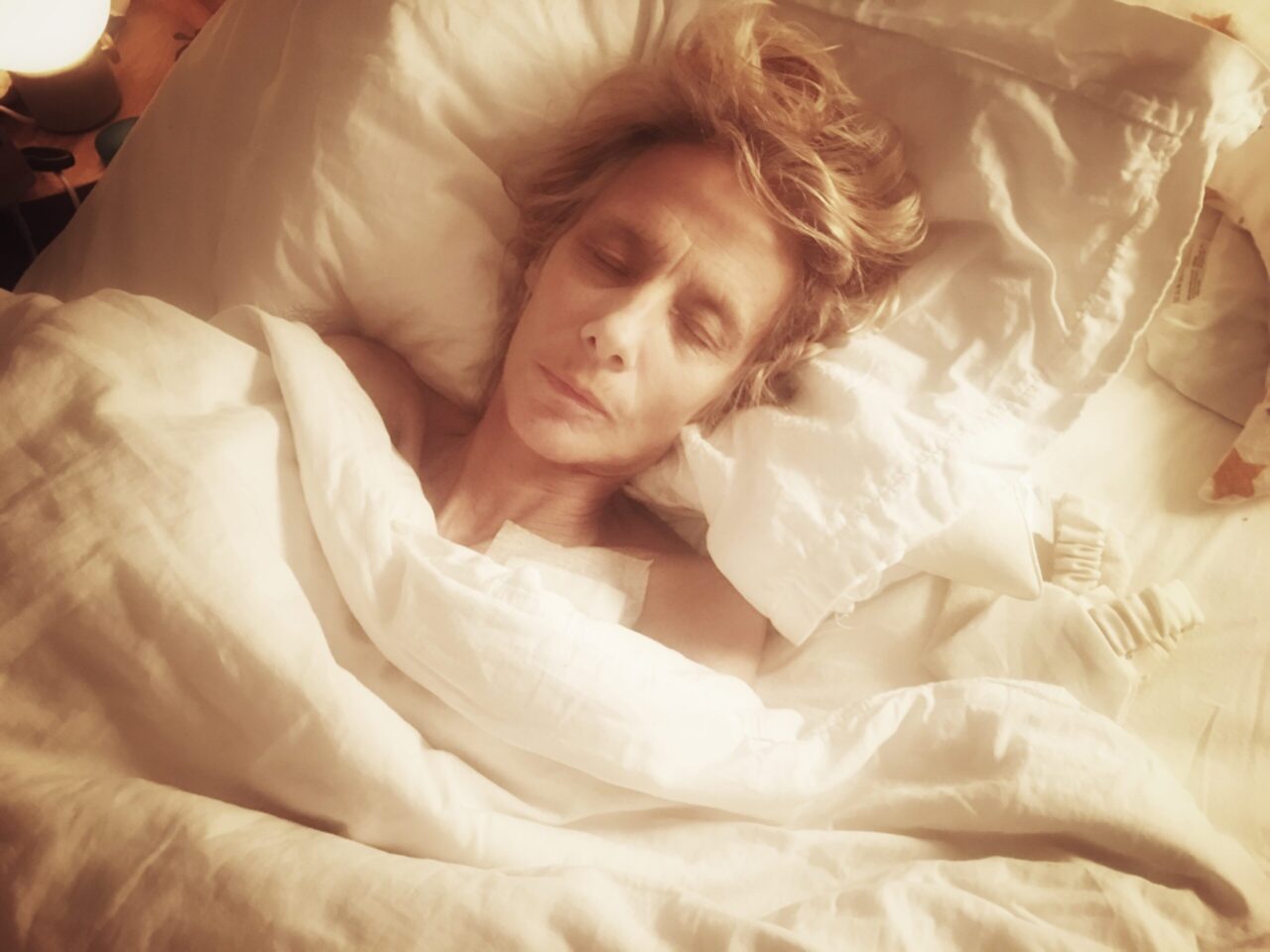 Eine Person schläft entspannt in einem hellen Bett, umgeben von weißen Laken und Kissen. Sanftes Licht umhüllt den Raum.