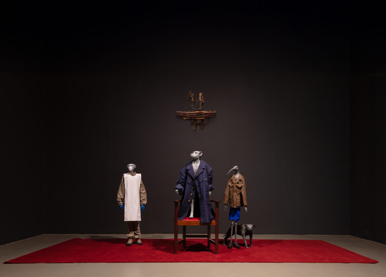 Exhibition with fashionable costumes on mannequins against a dark wall and red carpet, with a chair below.