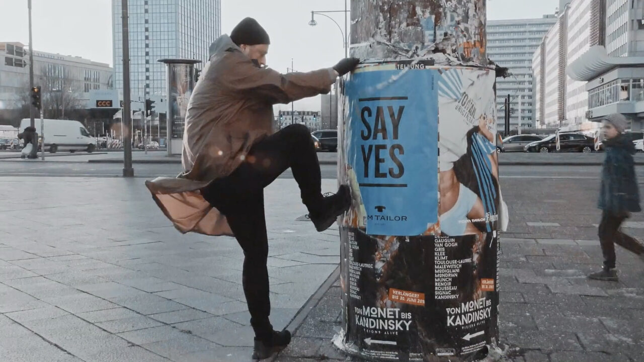 A man stands in front of a billboard that says "SAY YES" in an urban setting, with people in the background.