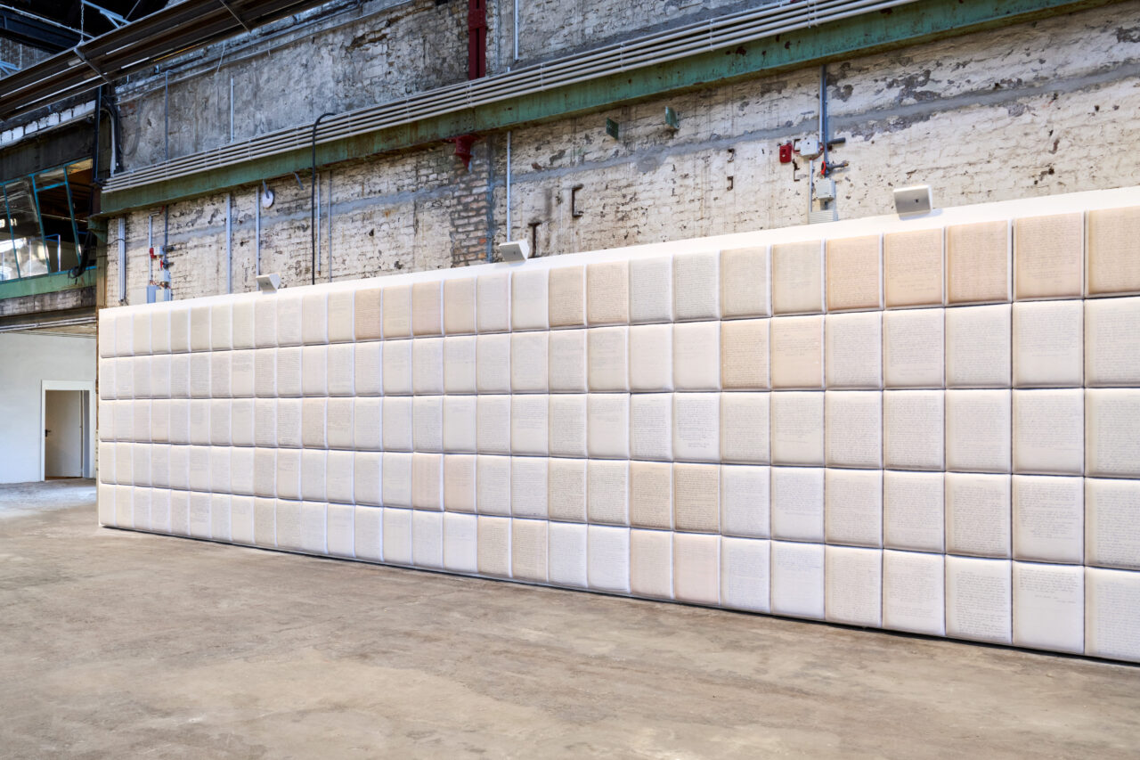 Wandinstallation mit quadratischen Tafeln, die mit handgeschriebenen Texten in einem minimalistischen Raum bedeckt sind.