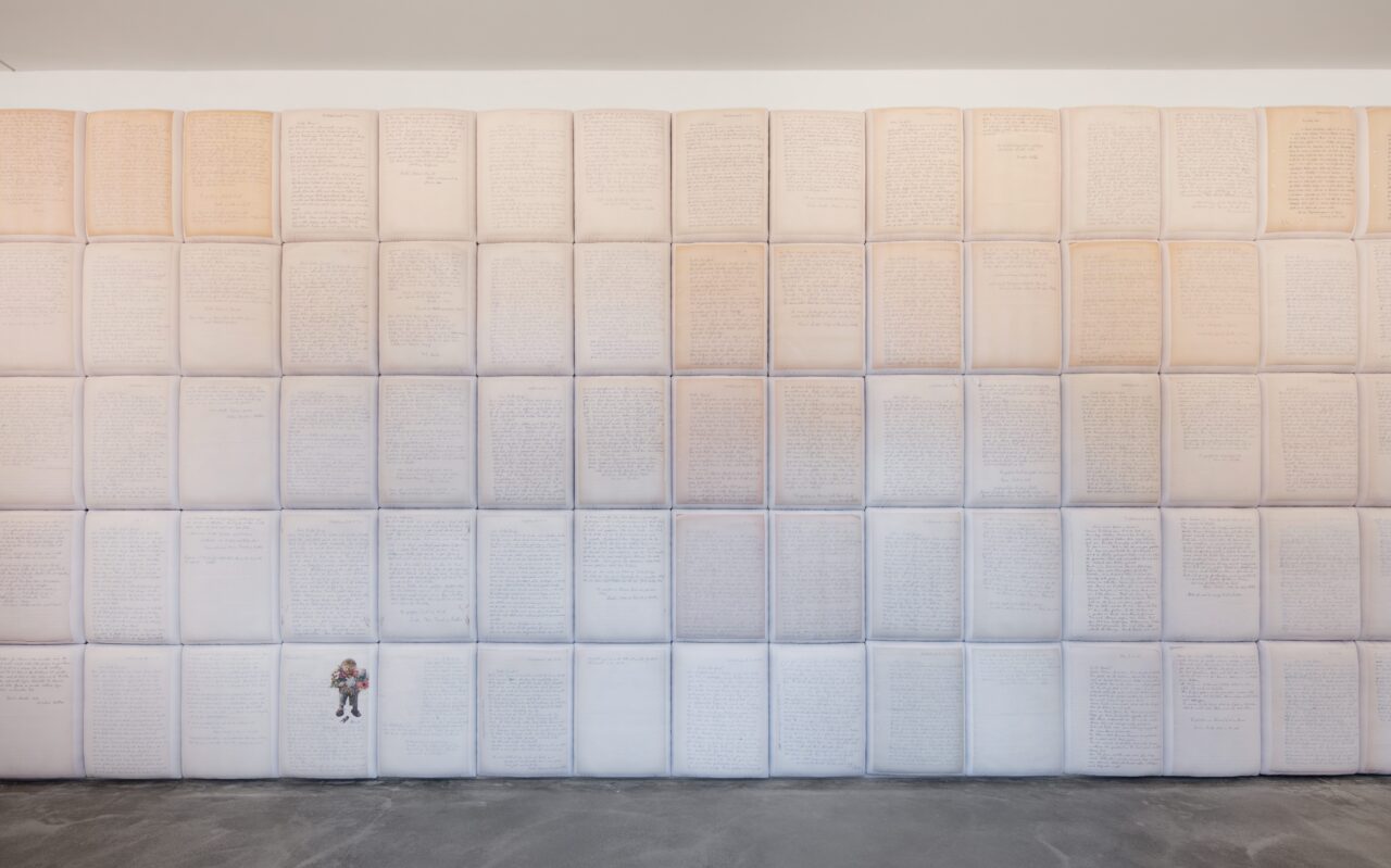 Eine Wand mit zahlreichen aufgeschlagenen Seiten, die handschriftlichen Text und eine kleine Illustrationsfigur zeigen.