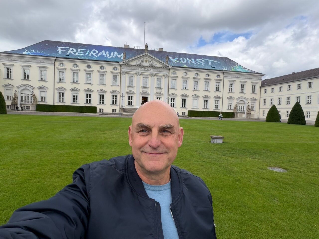 A person smiling in front of a large building with "FREIRAUM KUNST" painted on the roof, surrounded by greenery.