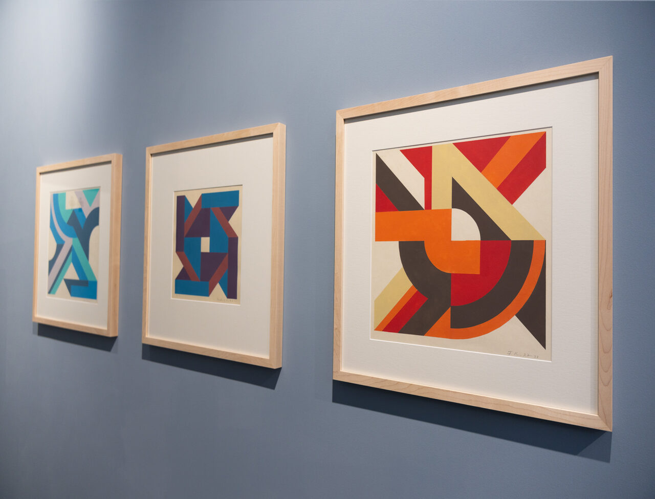 Three framed abstract artworks displayed on a blue wall, featuring geometric shapes and vibrant colors.