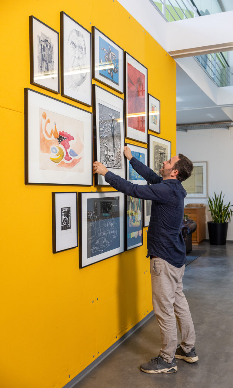 A person adjusting framed artworks on a bright yellow wall in a modern space.
