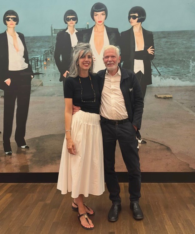 A couple poses in front of a picture featuring women in black suits at the beach. Stylish fashion, relaxed atmosphere.