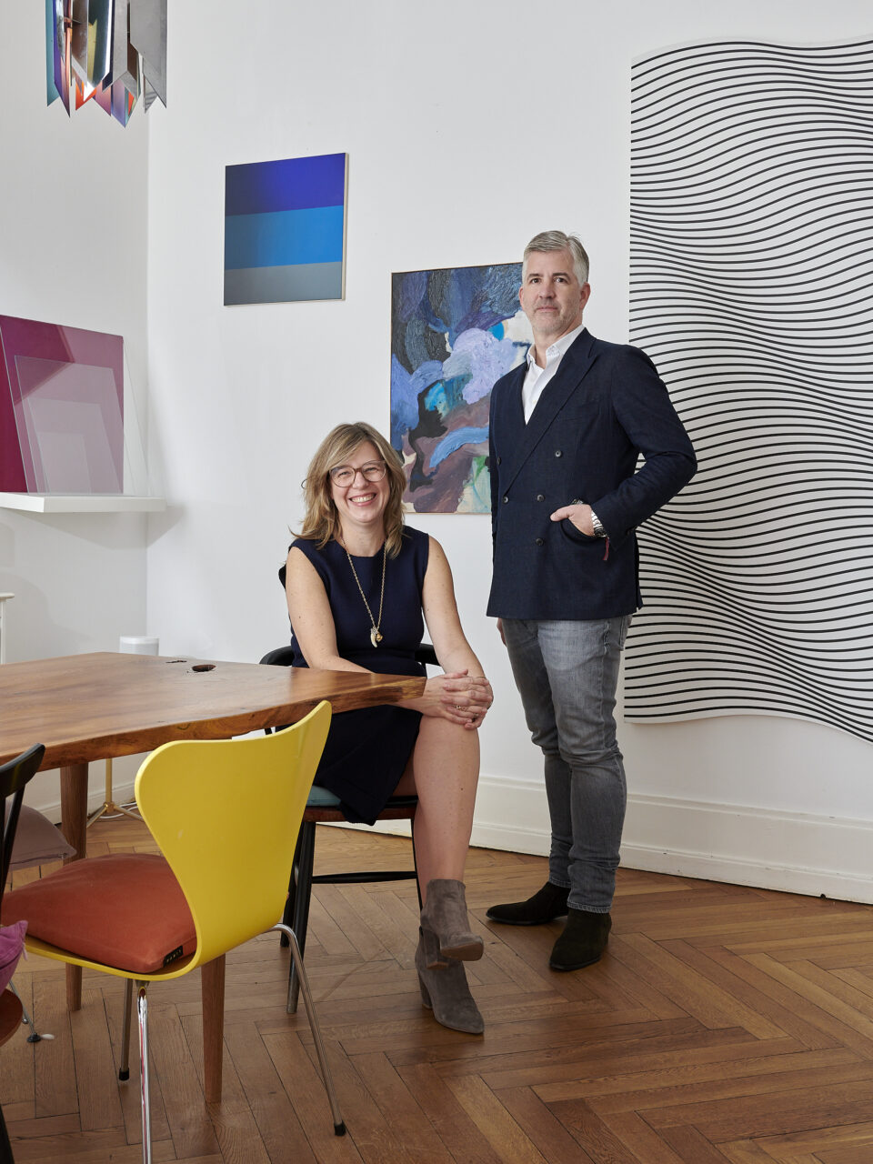 A man stands next to a seated woman in a modern, artistic room with colorful chairs and wall art.
