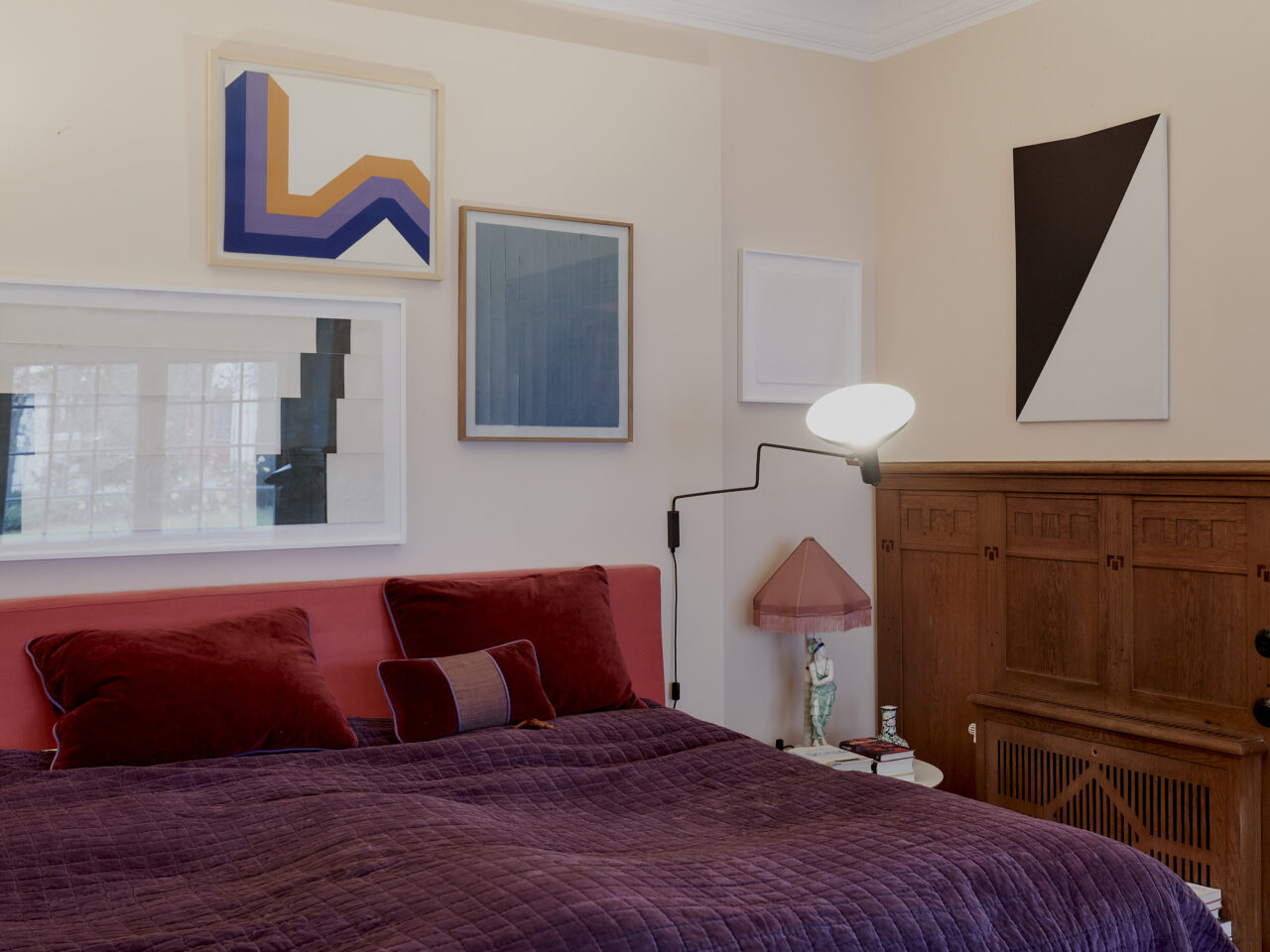 Modern bedroom with colorful artworks, a red bed, and stylish furniture in soft tones.