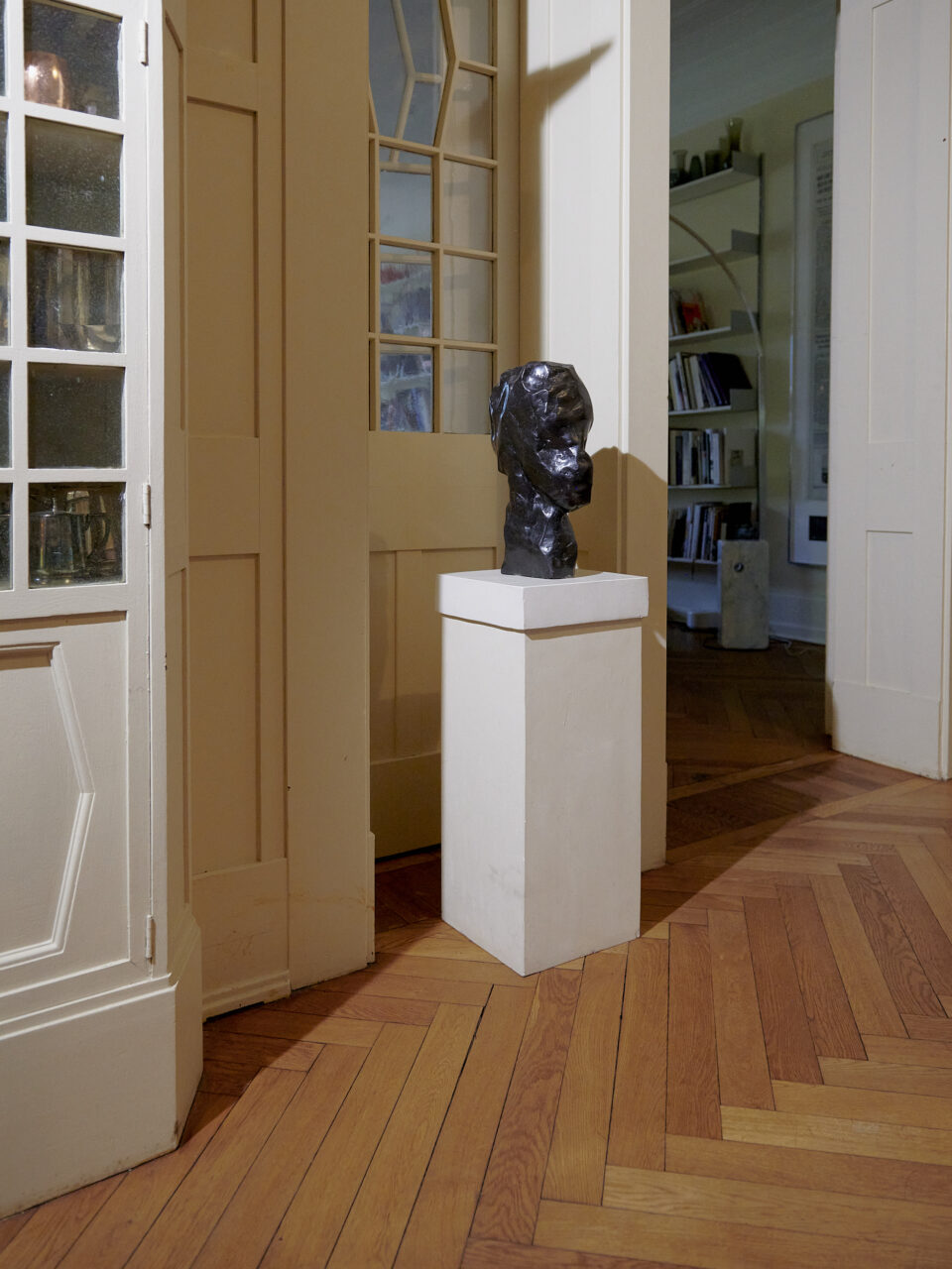 Sculpture of a head on a white pedestal in a room with wooden floors and glass panels.