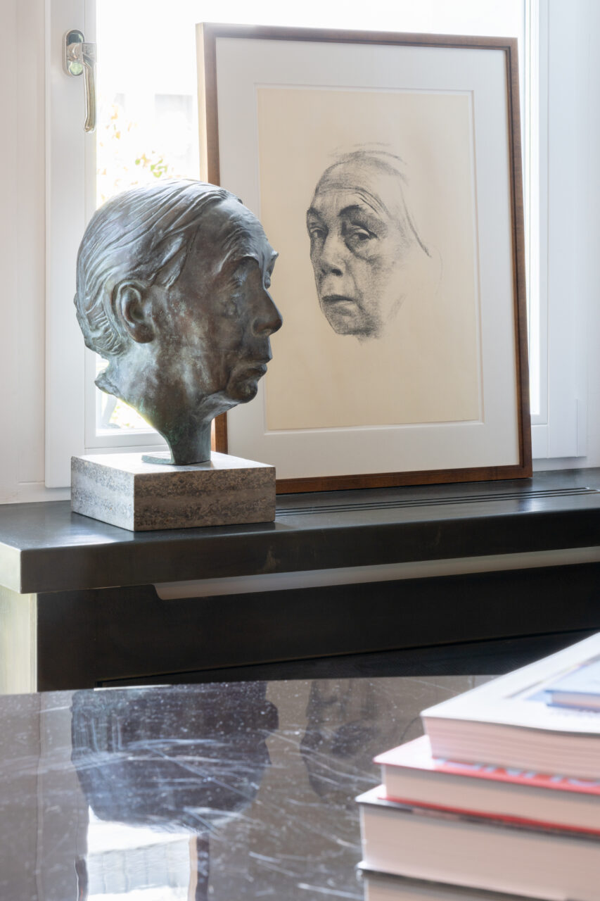 Bronze head and portrait drawing on a table, illuminated by window light. Stack of books in the foreground.
