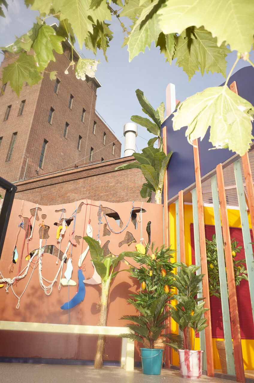 Bunte Wandgestaltung mit Pflanzen und strukturierten Elementen in einem urbanen Raum unter blauem Himmel.
